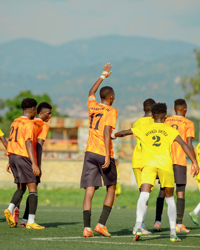 A youth soccer game with teams in orange and yellow jerseys takes place on a vibrant green field against a mountainous backdrop.